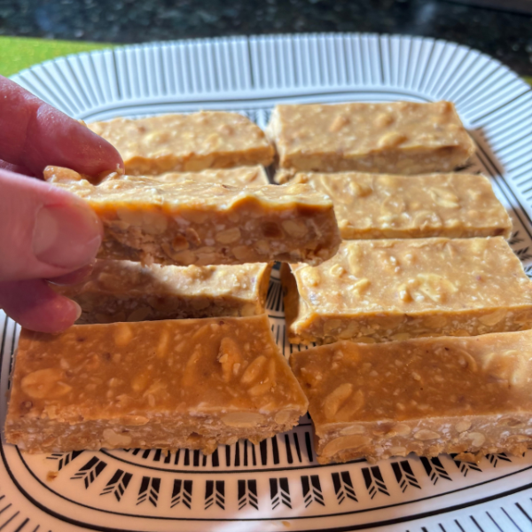 A hand holding up a no-bake peanut butter bar to show its texture and ingredients, with chopped peanuts and shredded coconut visible. More bars are arranged on a plate in the background.