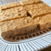 No-bake peanut butter bars cut into squares and arranged on a white plate, with visible bits of roasted peanuts and shredded coconut. The bars have a golden, slightly textured appearance and are set against a clean, natural background.