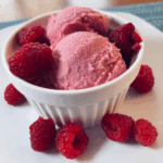 SCD raspberry frozen yogurt served in a white dish on a white plate, topped and surrounded by fresh raspberries.