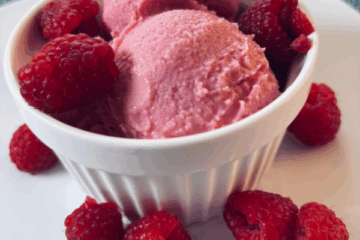SCD raspberry frozen yogurt served in a white dish on a white plate, topped and surrounded by fresh raspberries.