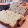 Sliced loaf of golden-brown red lentil bread on a wooden surface, with a soft, tender crumb and a bowl of red lentils in the background.